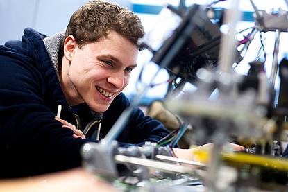 lachender junger Mann in dunkelblauem Ziphoodie an einer elektronischen Apparatur