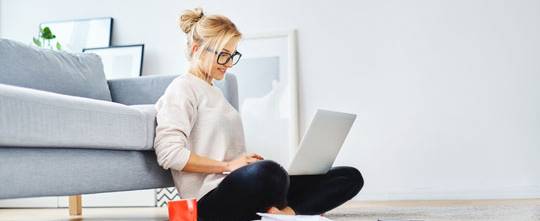 Young woman with glasses sits with laptop on her lap in front of a sofa