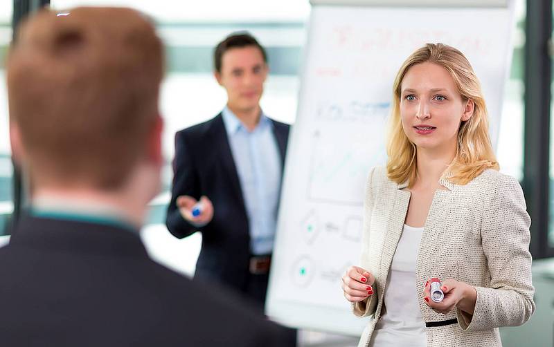 Zwei Personen stehen vor einer Flipchart und erklären einer dritten Person etwas. Die dritte Person steht im Vordergrund mit dem Rücken zur Kamera.