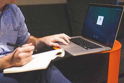 Eine Person, die mit einem Notizblock und Stift in der Hand, auf einem schwarzen Sofa sitzt. Neben der Person befindet sich kleiner Tisch, auf dem ein Laptop ist und die Person scrollt zu einem Feld, welches am Laptop Bildschirm zu sehen ist.