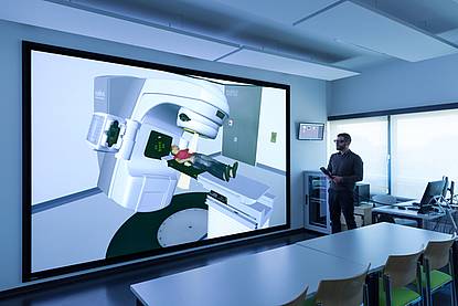 Klassenraum mit einer großen Leinwand, auf der eine virtuelle und lebensgroße Simulation von der Strahlentherapie bei einer Person zu sehen ist.