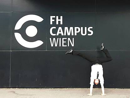 Mann in weißen Hemd, Krawatte und Anzugshose vor einer Wand, auf der das F H Campus Wien Logo angebracht ist, welches aus dem künstlerisch gezeichnete C-Form und daneben der Schriftzug FH Campus Wien besteht. Der Mann macht gerade einen Handstand und formt dabei mit seinen Füssen ein Häkchen.