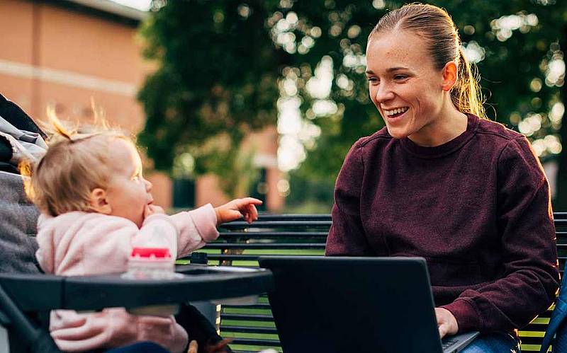 Junge Frau auf einer Parkbank mit Laptop auf dem Schoß. Neben ihr ihr Kleinkind im Kinderwagen. 