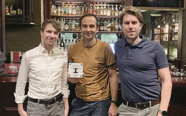 Drei Männer stehen vor einer Bar, der mittlere hält die gemeinsam gewonnene Preistrophäe in der Hand