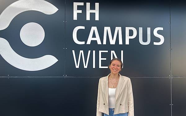 Eine junge Frau steht vor einer Wand auf der FH Campus Wien zu lesen ist