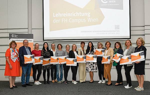 Personen stehen vor einer Leinwand, auf der Lehreinrichtung der FH Campus Wien zu lesen ist. Jede Person hält eine orange Lehrtafel in der Hand.