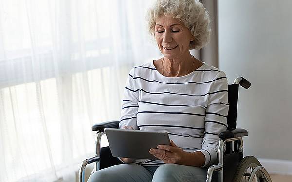 eine ältere Frau sitzt im Rollstuhl und hat ein Tablet in der Hand