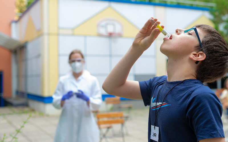 Kind beim Gurgel der speziellen Salzlösung im Freien, im Hintergrund stoppt die Studierende die Zeit