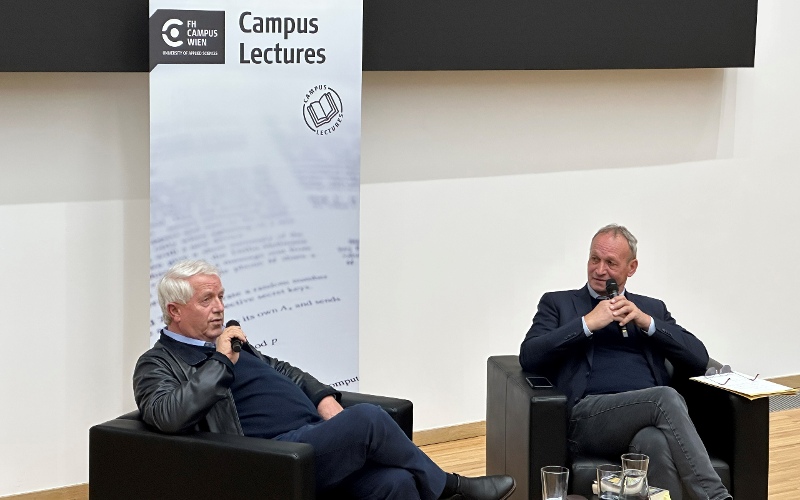 Zwei Männer sitzen auf Ledersesseln und halten ein Mikrophon in der Hand, im Hintergrund ist ein Roll-up zu sehen auf dem Campus Lectures steht