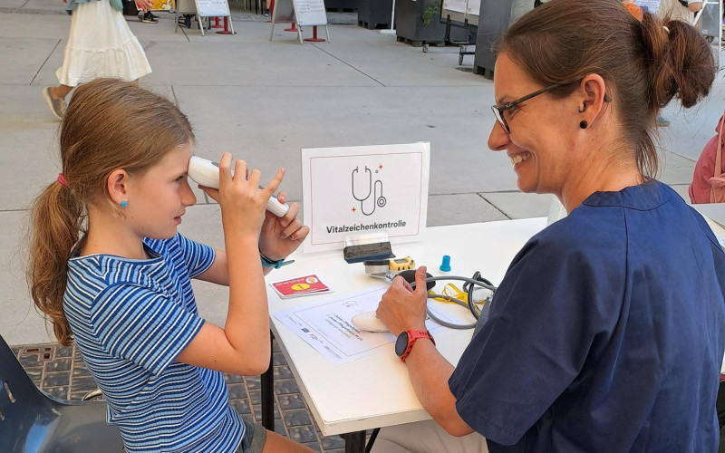 ein kind hält sich ein Fieberthermometer an die Stirn, daneben sitzt eine Frau