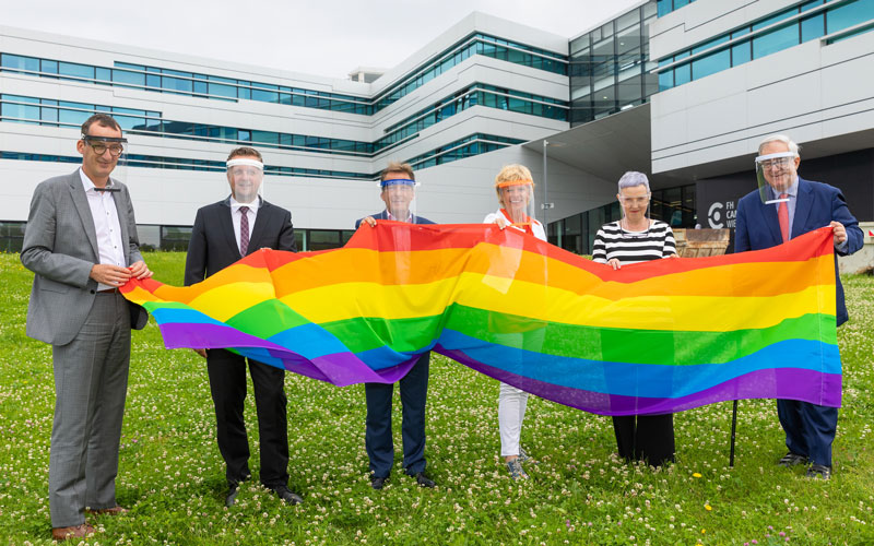 Mitarbeiterinnen und Mitarbeiter halten die Regenbogenfahne vor dem F H Campus Wien Gebäude hoch