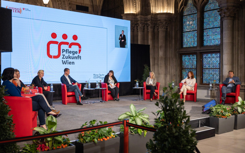 Acht Expert*innen sitzen in einer Diskussionsrunde auf der Bühne in der Volkshalle, Wiener Rathaus