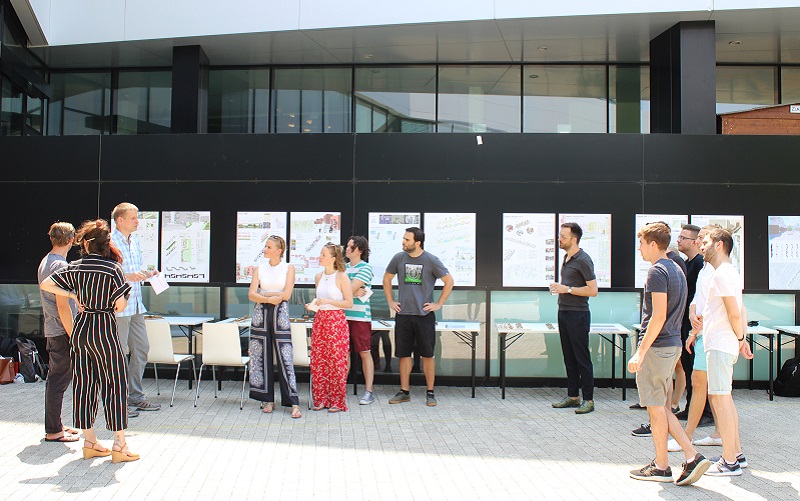 Frauen und Männer vor an Pinnwändwn montierten Plänen, Plakaten und Modellen bei der Entwurfspräsentation