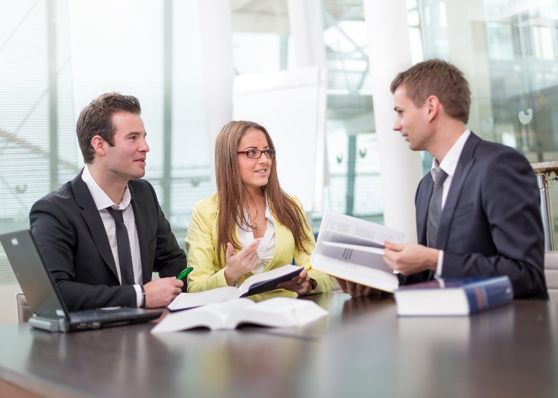 Drei Personen, ein Mann, eine Frau und ein Mann, alle gekleidet in Businesskleidung sitzen an einem Tisch. Vor ihnen liegen aufgeschlagene Bücher. Sie unterhalten sich miteinander.