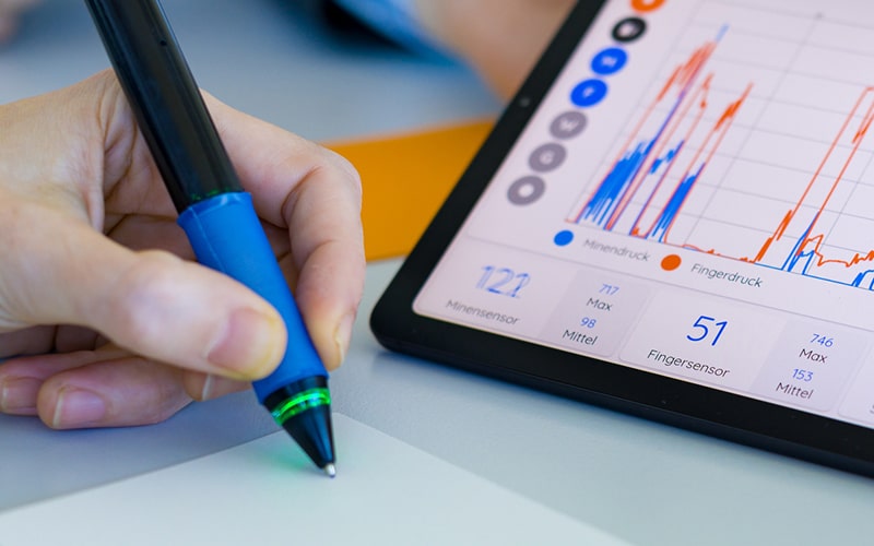 Nahaufnahme einer Hand mit Kugelschreiber beim Notieren von Gesundheitsdaten, die auf einem Tablet in Rot und Blau dargestellt sind