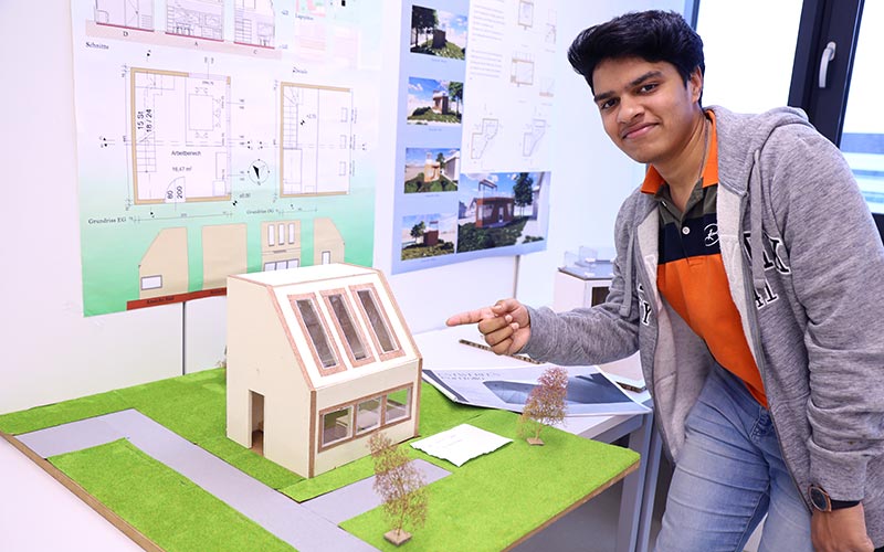 Studierender vor selbstentworfenen Plänen und einem Modell eines Arbeitsraumes im Grünen